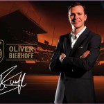 Mann im Anzug steht vor der Kulisse eines Fußballstadions mit Deutschland-Branding-Elementen und einem Namensschild mit der Aufschrift „Oliver Bierhoff“. Fritten, Fussball & Bier - www.soccer-warriors.de