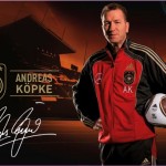 Ein Mann in der Kleidung eines Fußballtrainers posiert mit einem Fußball, auf dem Logos von adidas und dem Deutschen Fußballverband zu sehen sind, darunter eine Unterschrift. Fritten, Fussball & Bier - www.soccer-warriors.de