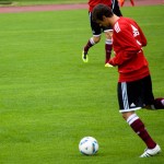 Ein Fußballspieler im roten Trikot, der während eines Testspiels auf einem grünen Spielfeld einen Ball kicken will. Fritten, Fussball & Bier - www.soccer-warriors.de
