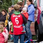 Eine Frau mit einem Kind steht inmitten einer Menschengruppe im Freien, im Vordergrund ein Kind im roten „1. FC Nürnberg Testspiel“-Trikot. Fritten, Fussball & Bier - www.soccer-warriors.de