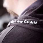 Nahaufnahme eines schwarzen Kleidungsstücks mit dem in Weiß gestickten deutschen Text „wir sind der glubb“ für den 1. FC Nürnberg. Fritten, Fussball & Bier - www.soccer-warriors.de