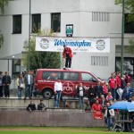 Zuschauer versammeln sich bei einer Sportveranstaltung unter Transparenten und Schildern für ein Testspiel um einen roten Transporter. Fritten, Fussball & Bier - www.soccer-warriors.de