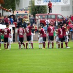 Eine Fußballmannschaft in kastanienbraunen Uniformen drängt sich vor einem Testspiel auf einem Feld zusammen, im Hintergrund Zuschauer. Fritten, Fussball & Bier - www.soccer-warriors.de