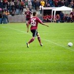 Ein Fußballspieler in burgunderfarbener Uniform kickt während eines Testspiels mit Zuschauern im Hintergrund einen Ball. Fritten, Fussball & Bier - www.soccer-warriors.de
