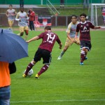Auf einem nassen Feld läuft ein Fußballspiel mit Spielern des 1. FC Nürnberg und Racing Santander in kastanienbraunen und gelben Trikots, während Zuschauer unter Sonnenschirmen zuschauen. Fritten, Fussball & Bier - www.soccer-warriors.de