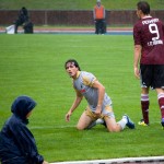 Ein Fußballspieler kniet auf einem nassen Spielfeld und blickt bei Regen zur Seitenlinie, mit anderen Spielern und einem Zuschauer im Hintergrund während eines Testspiels. Fritten, Fussball & Bier - www.soccer-warriors.de