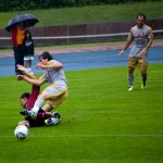 Fußballspieler auf einem nassen Feld, einer rutscht nach dem Ball, während ein anderer und ein Zuschauer mit Regenschirm das Testspiel zwischen Racing Santander und dem 1. FC Nürnberg verfolgen. Fritten, Fussball & Bier - www.soccer-warriors.de