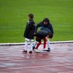 Drei Kinder auf einem nassen Leichtathletikplatz, eines bückt sich, um neben einer Fahne mit dem Wappen des FC Nürnberg etwas aufzuheben. Fritten, Fussball & Bier - www.soccer-warriors.de