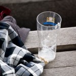 Ein leeres Pintglas mit Schaumresten steht auf einer Holzbank neben einem drapierten karierten Mantel und einem roten Kleidungsstück von Racing Santander. Fritten, Fussball & Bier - www.soccer-warriors.de