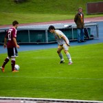 Zwei Fußballspieler auf dem Feld während eines Testspiels, einer bereitet sich darauf vor, den Ball zu schießen, während Zuschauer hinter einer Absperrung zuschauen. Fritten, Fussball & Bier - www.soccer-warriors.de
