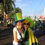 Zwei Personen lächeln und umarmen sich, tragen leuchtend grüne und gelbe Hüte und Accessoires, möglicherweise bei einer Straßenfeier oder einer Sportveranstaltung in Brasilien. Fritten, Fussball & Bier - www.soccer-warriors.de