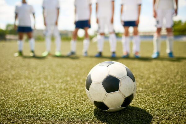 Ein Fußball liegt auf einem Kunstrasen. Im unscharfen Hintergrund stehen fünf Spieler in weißen Trikots und Shorts. Fritten, Fussball & Bier - www.soccer-warriors.de